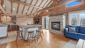A spacious log cabin interior with a stone fireplace, wooden bar stools, and a blue sofa by a large window.