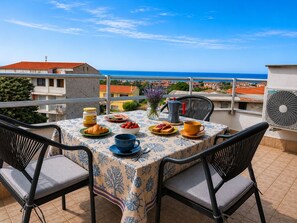 Une terrasse avec une table dressée pour le petit-déjeuner, des chaises, une vue sur la mer et un bâtiment en arrière-plan.