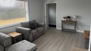 A modern living room with a grey sofa, a small wooden table, and a console table with decorative items. - 3 Bed Cottage - Sleeps 5 - Pets - Hottub - Garden (King's Lynn)