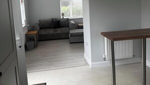 A modern living room with a grey sofa, a wooden coffee table, and a view of the outside through a window with blinds. - 3 Bed Cottage - Sleeps 5 - Pets - Hottub - Garden (King's Lynn)