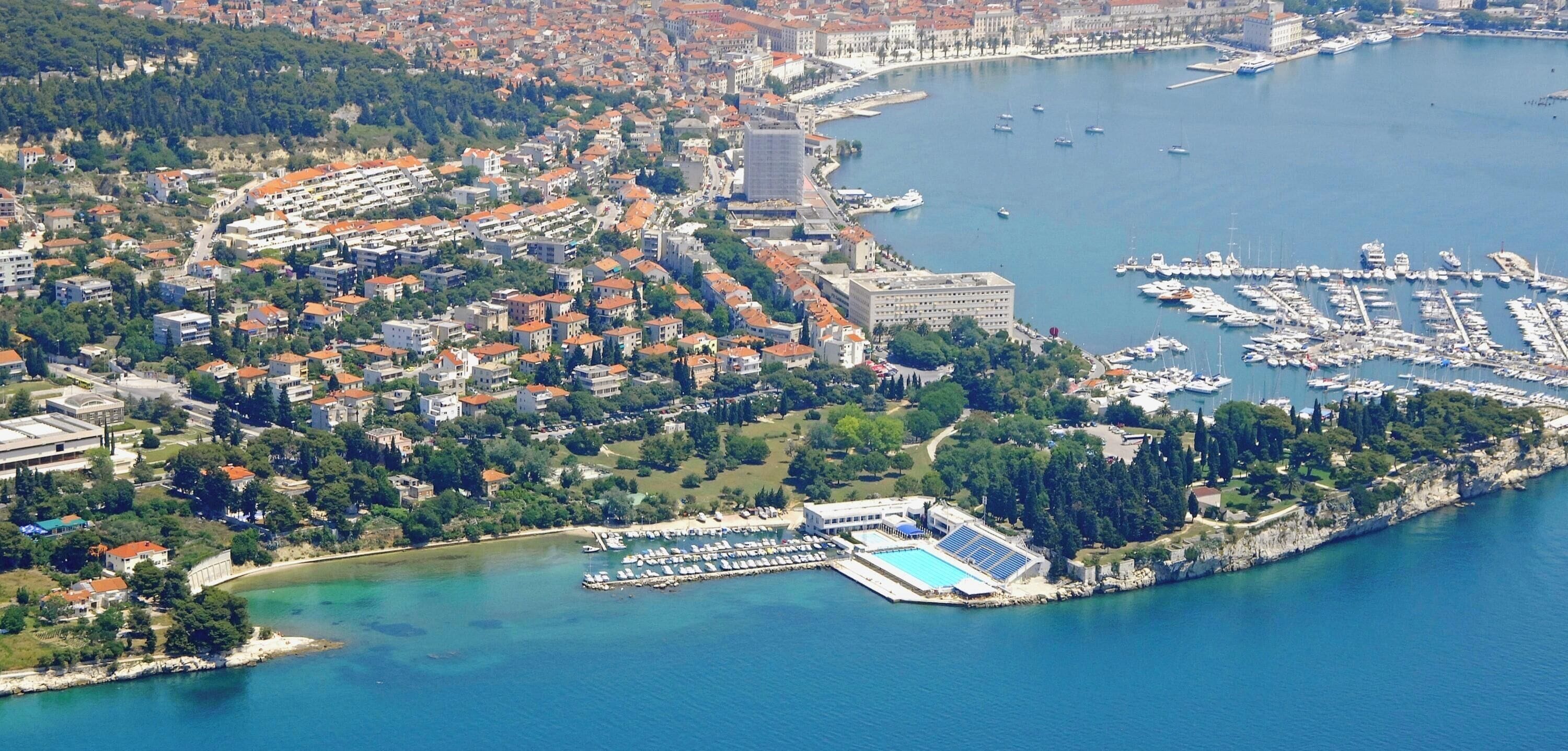 An aerial view of a coastal town with a marina, residential buildings, and a swimming pool complex.