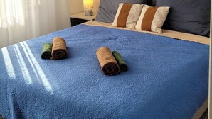 A hotel room with a blue quilted bedspread, two rolled towels, a nightstand with a lamp, a painting of palm trees, and a window with white curtains. - Charming apt With Balcony in the Heart of Split (Split)