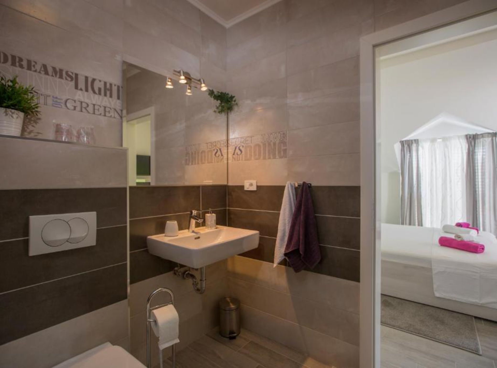 Une salle de bain moderne avec un grand miroir, un lavabo et des toilettes. On y trouve un porte-serviettes avec des serviettes, une poubelle et une plante accrochée au mur.