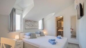 A bedroom with a bed, bedside tables, a desk, a chair, a window, a painting, and a bathroom visible through an open door. - Adriatic Pearl (Solta)