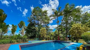 A swimming pool with a blue and red border, surrounded by tropical vegetation and a clear blue sky. - Cabinas Faro Arenal (La Fortuna)