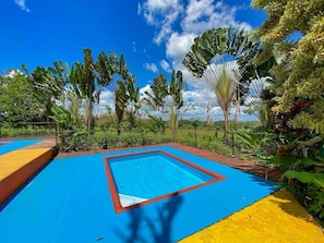 A pool area with a blue and yellow deck, surrounded by tropical plants and trees. - Cabinas Faro Arenal (La Fortuna)