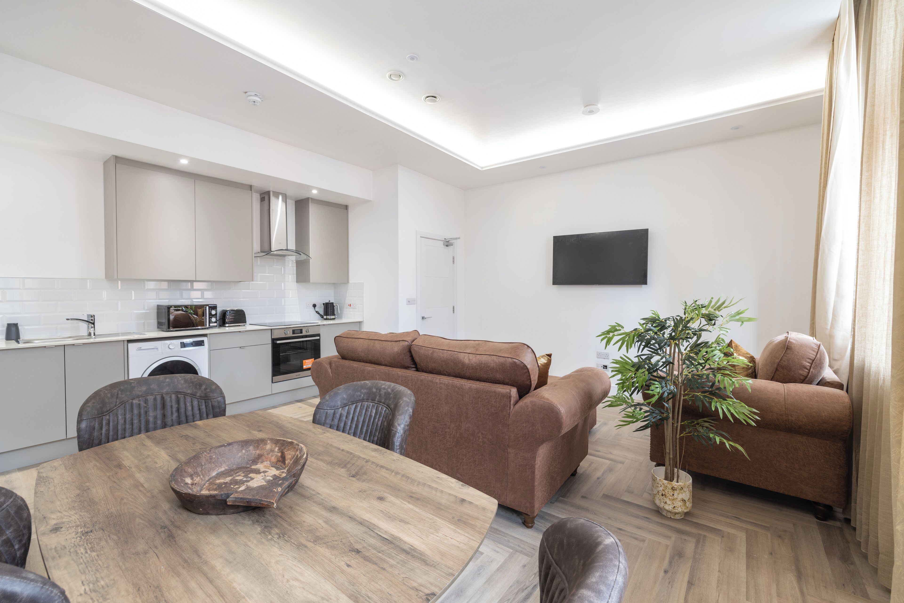 A modern kitchen with built-in appliances, a dining area with a wooden table, and a living room with a sofa and a potted plant.