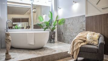 A modern bathroom with a freestanding bathtub, a small Buddha statue, a potted plant, and a grey sofa with a throw blanket.