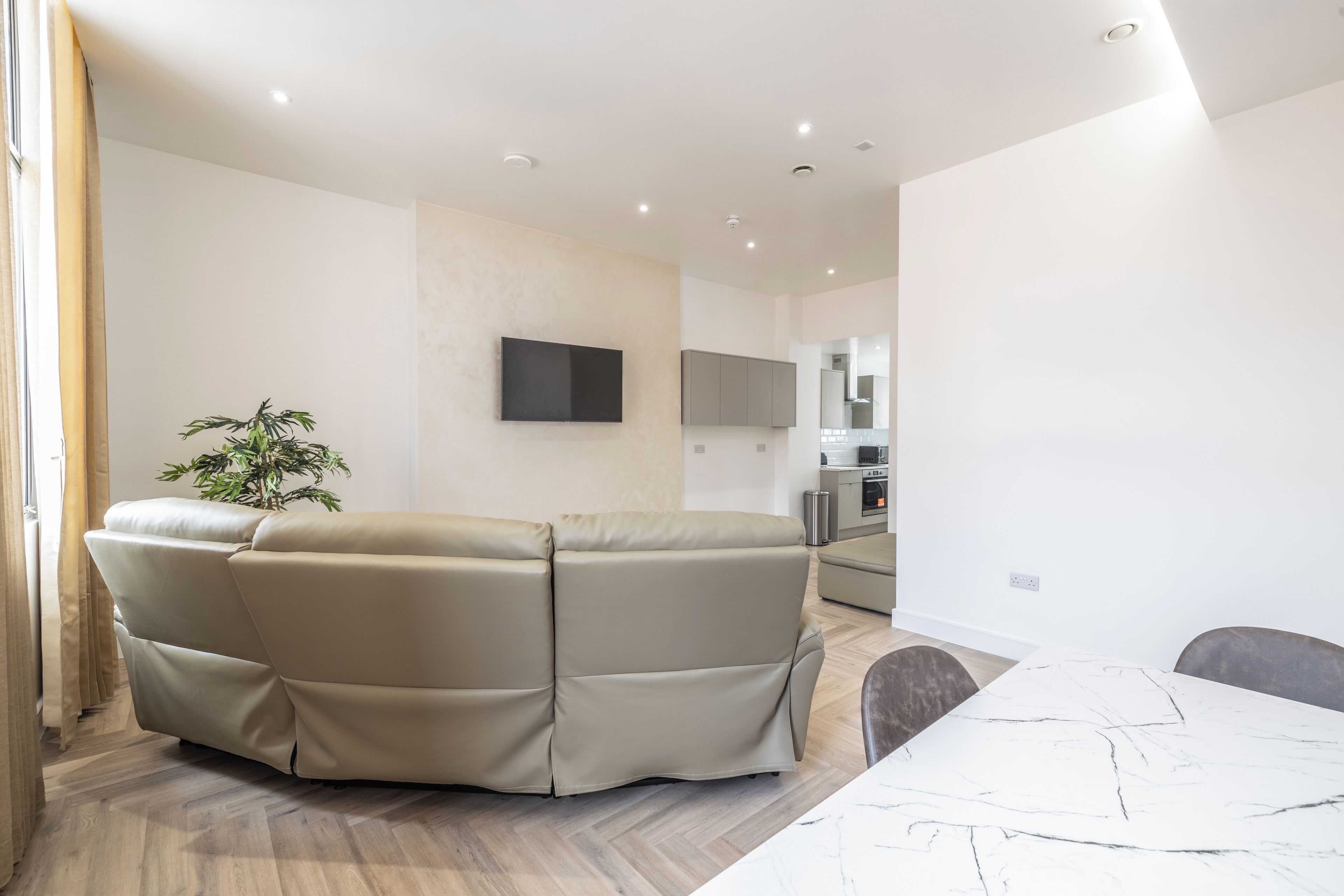 Una sala de estar moderna con un sofá seccional color beige, una mesa de comedor con sillas, un televisor de pantalla plana montado en la pared y una zona de cocina al fondo.