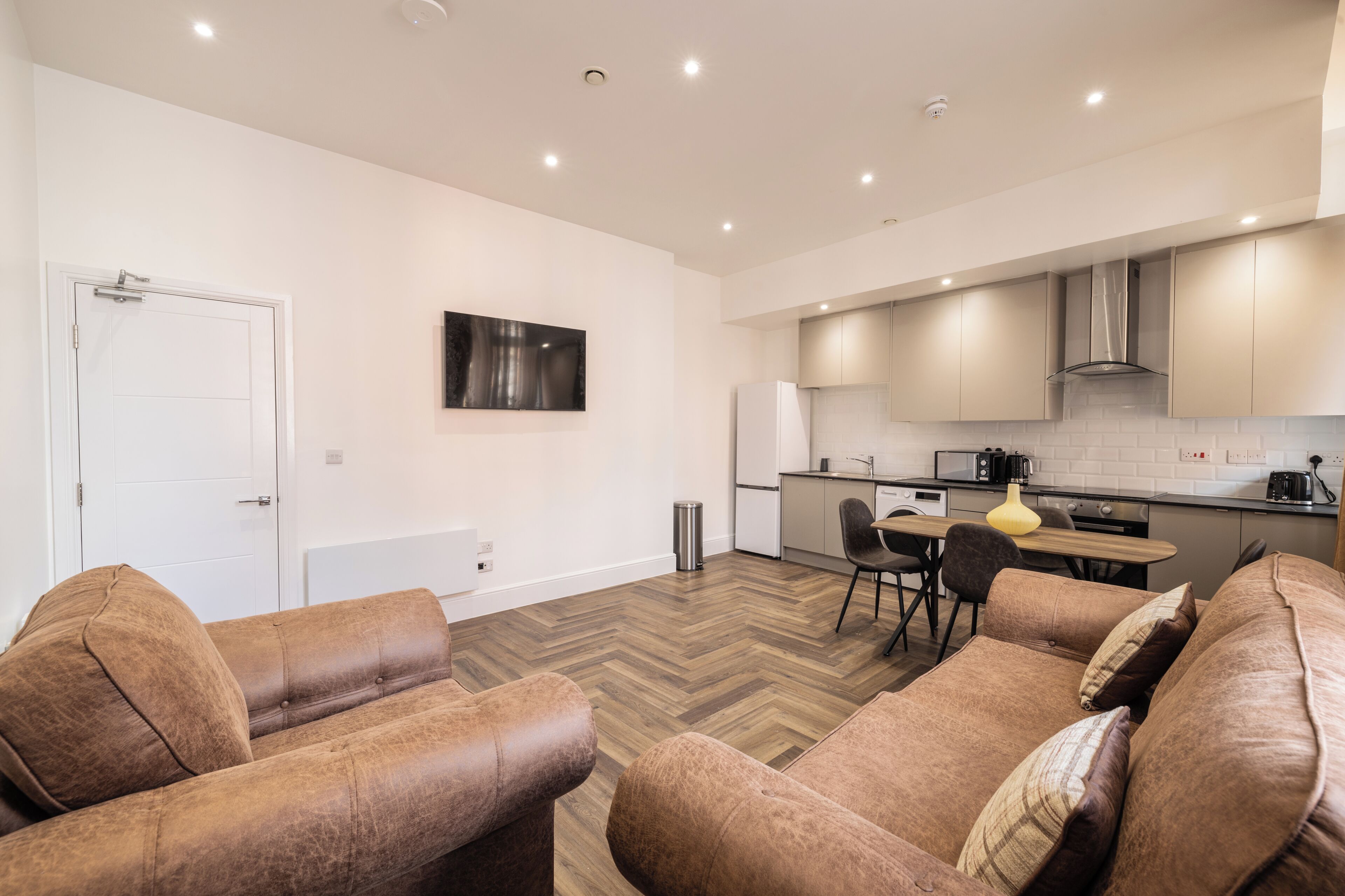 A modern living room with a sofa set, a dining table with chairs, a kitchen area with cabinets and appliances, and a white door.