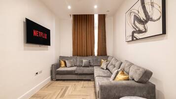 A modern living room with a gray sectional sofa, a mounted TV displaying 'NETFLIX,' and a large abstract painting on the wall.