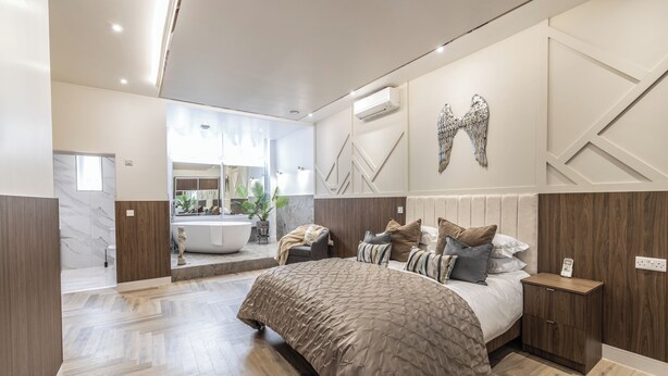 Un dormitorio moderno con una cama grande, una mesita de noche, un espejo y un baño a la vista en el fondo.