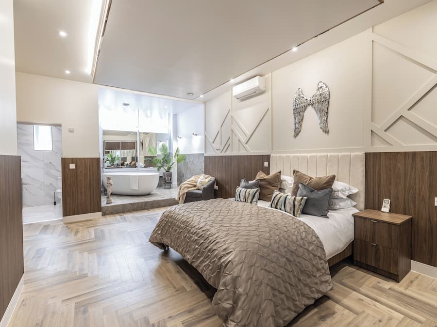 Quarto moderno com cama grande, mesa de cabeceira, espelho e banheiro visível ao fundo.