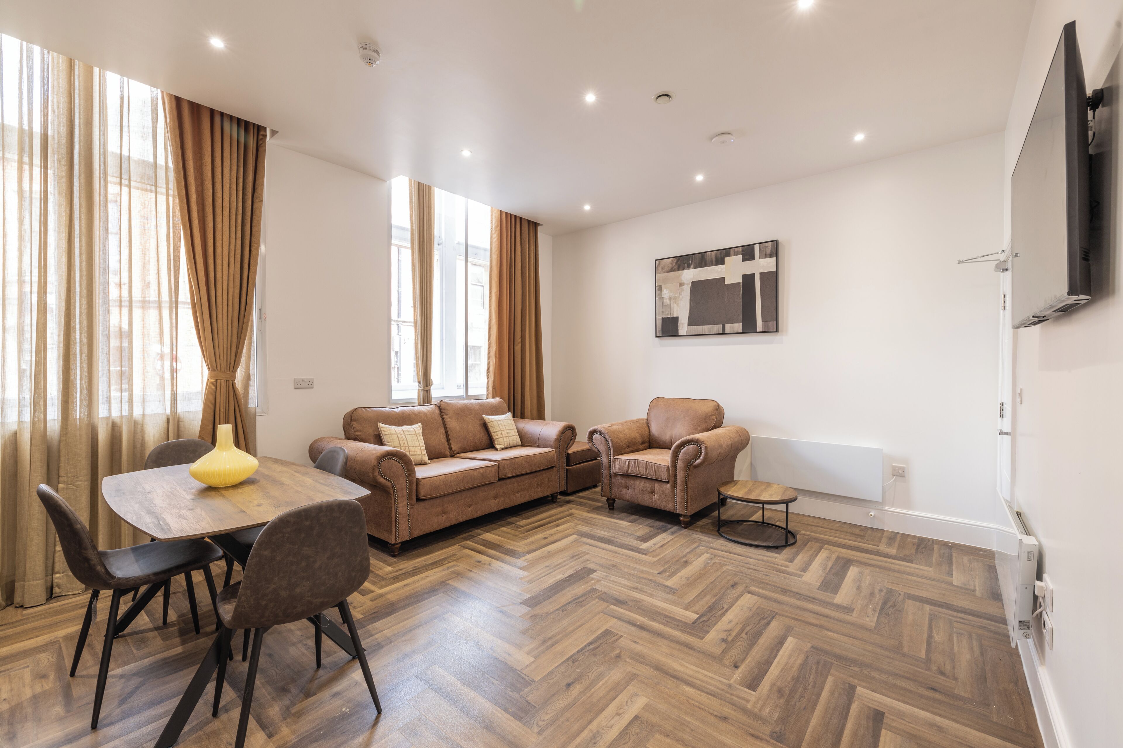 A modern living room with a wooden herringbone floor, a brown leather sofa set, a dining table with chairs, and a flat-screen TV mounted on the wall.