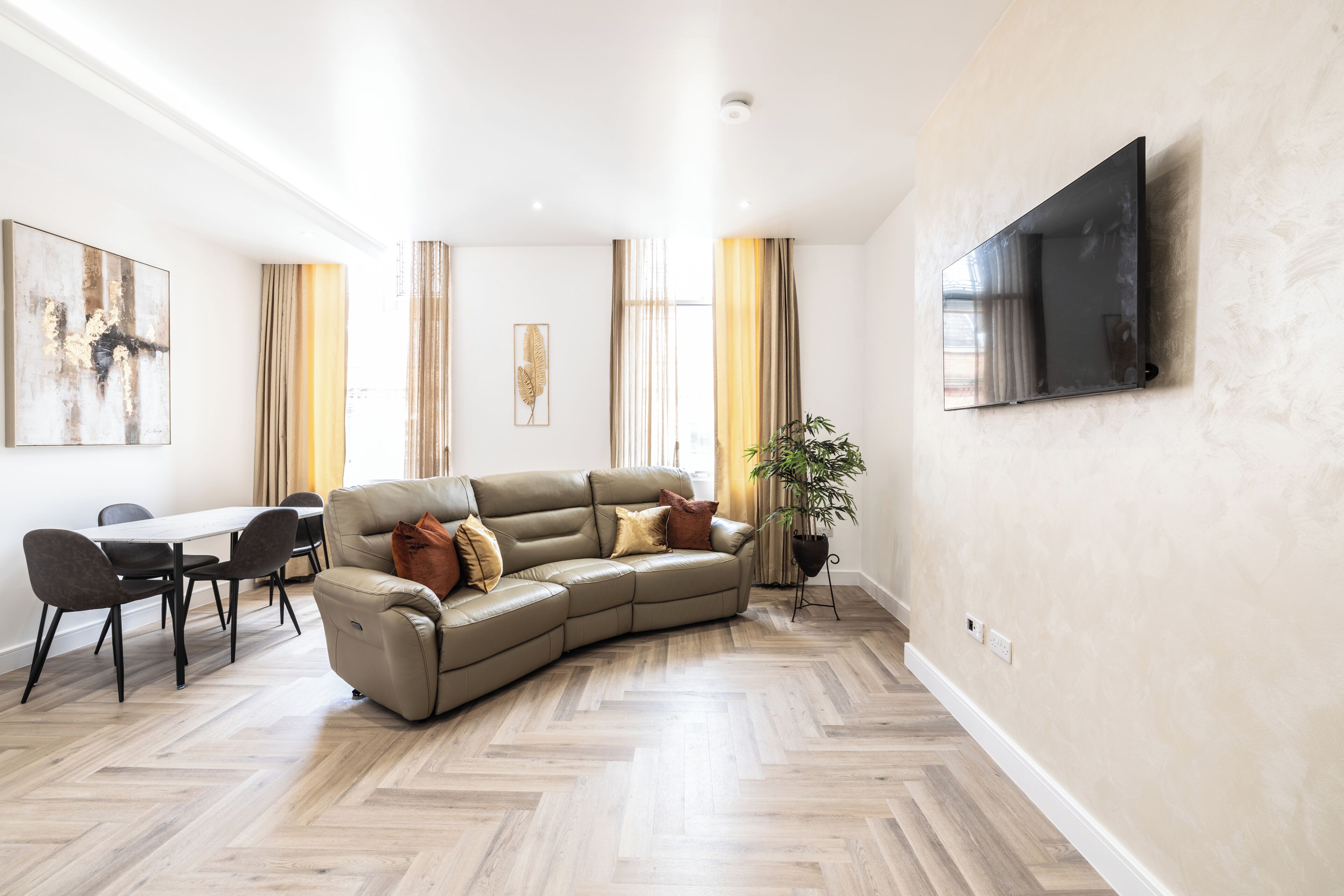 Een moderne woonkamer met een hoekbank, een eettafel met stoelen, een flatscreen televisie aan de muur en een potplant.