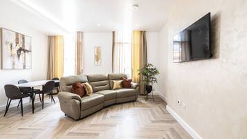 Een moderne woonkamer met een hoekbank, een eettafel met stoelen, een flatscreen televisie aan de muur en een potplant.