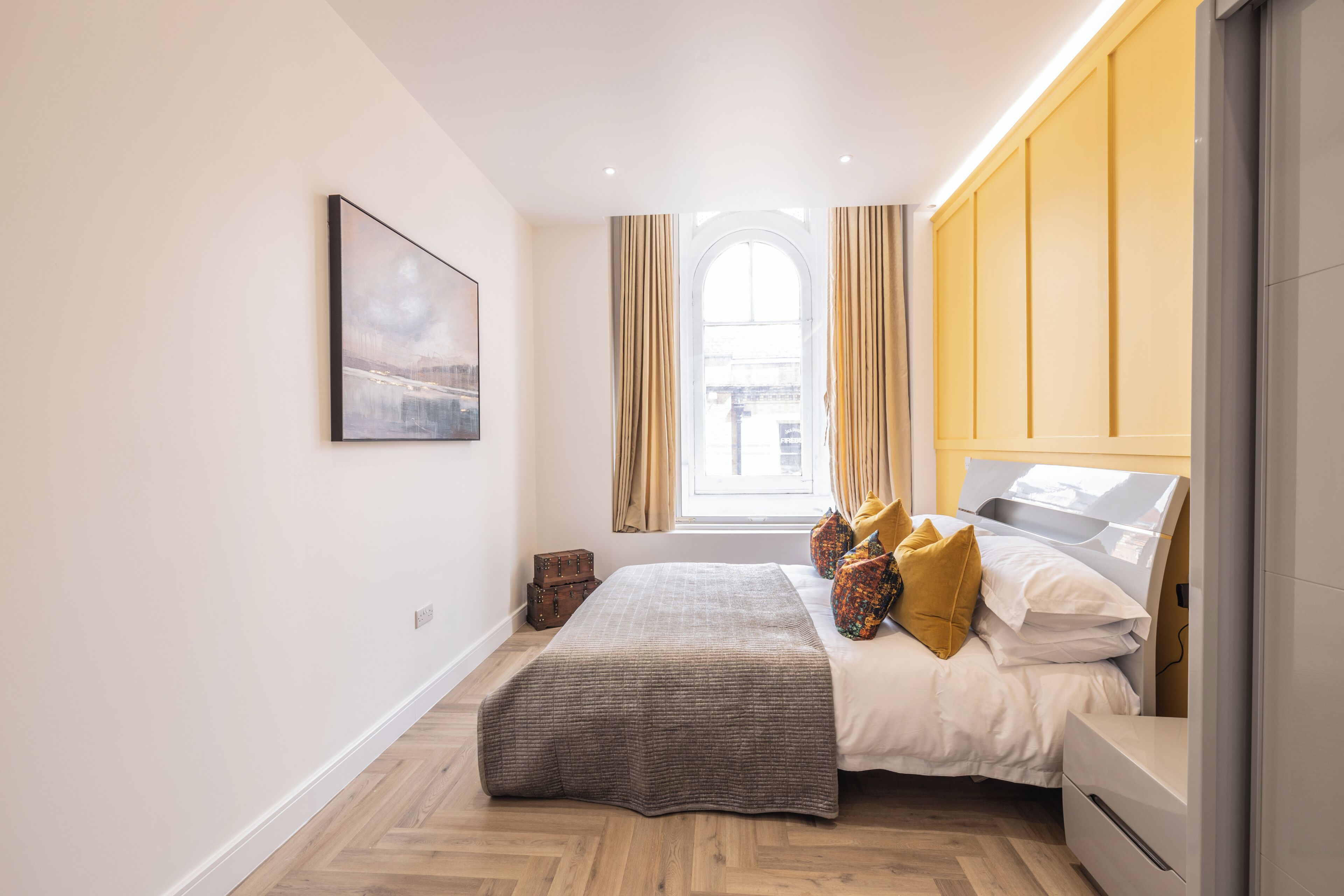 A bedroom with a bed, a bedside table, a window with curtains, a picture on the wall, and a herringbone floor pattern.