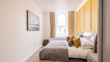 A bedroom with a bed, a bedside table, a window with curtains, a picture on the wall, and a herringbone floor pattern.