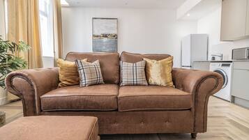 A brown leather sofa with plaid cushions in a modern living room with a kitchen in the background.