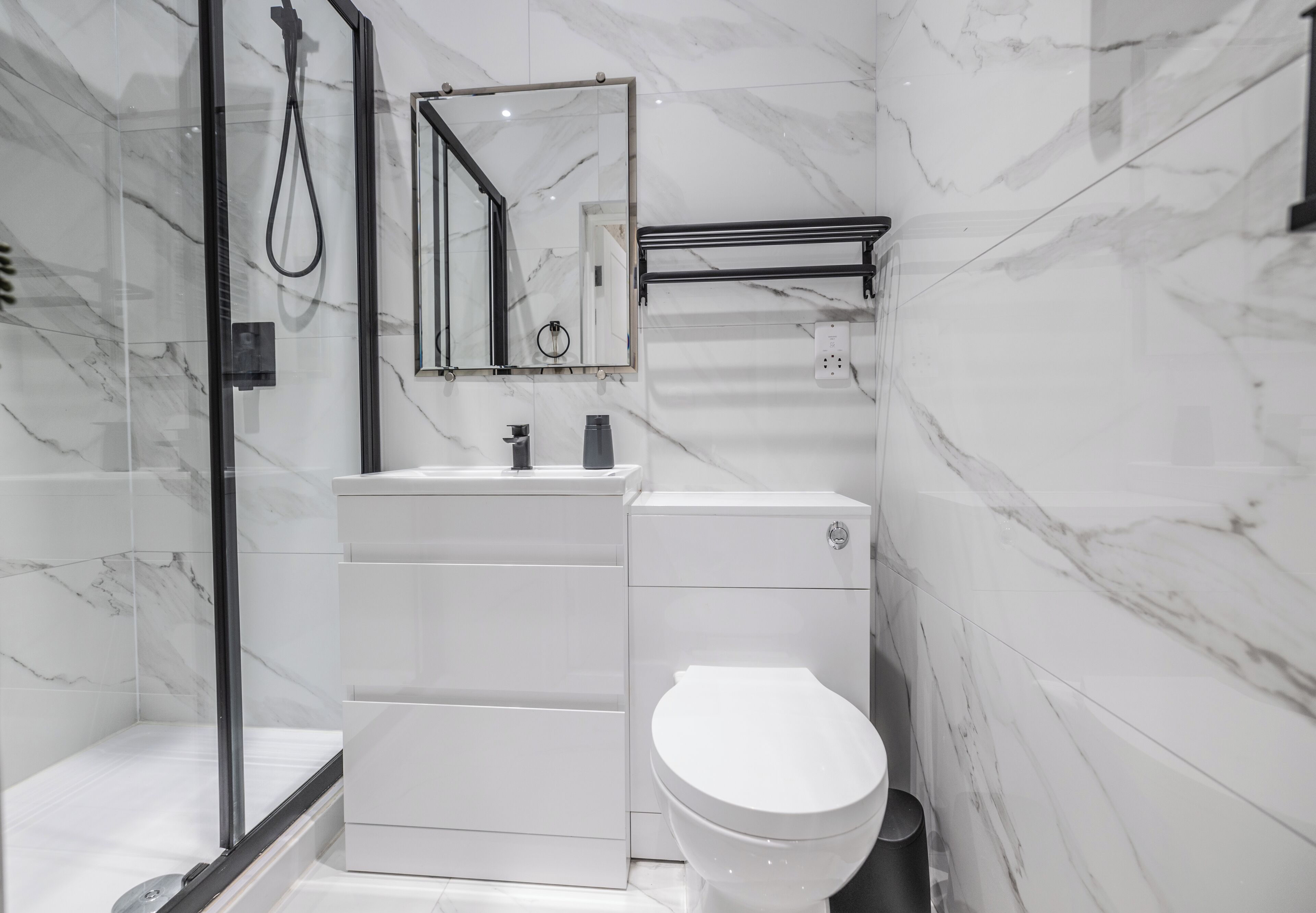 Un baño moderno con cabina de ducha de vidrio, inodoro blanco, lavamanos con espejo y un perchero para toallas.