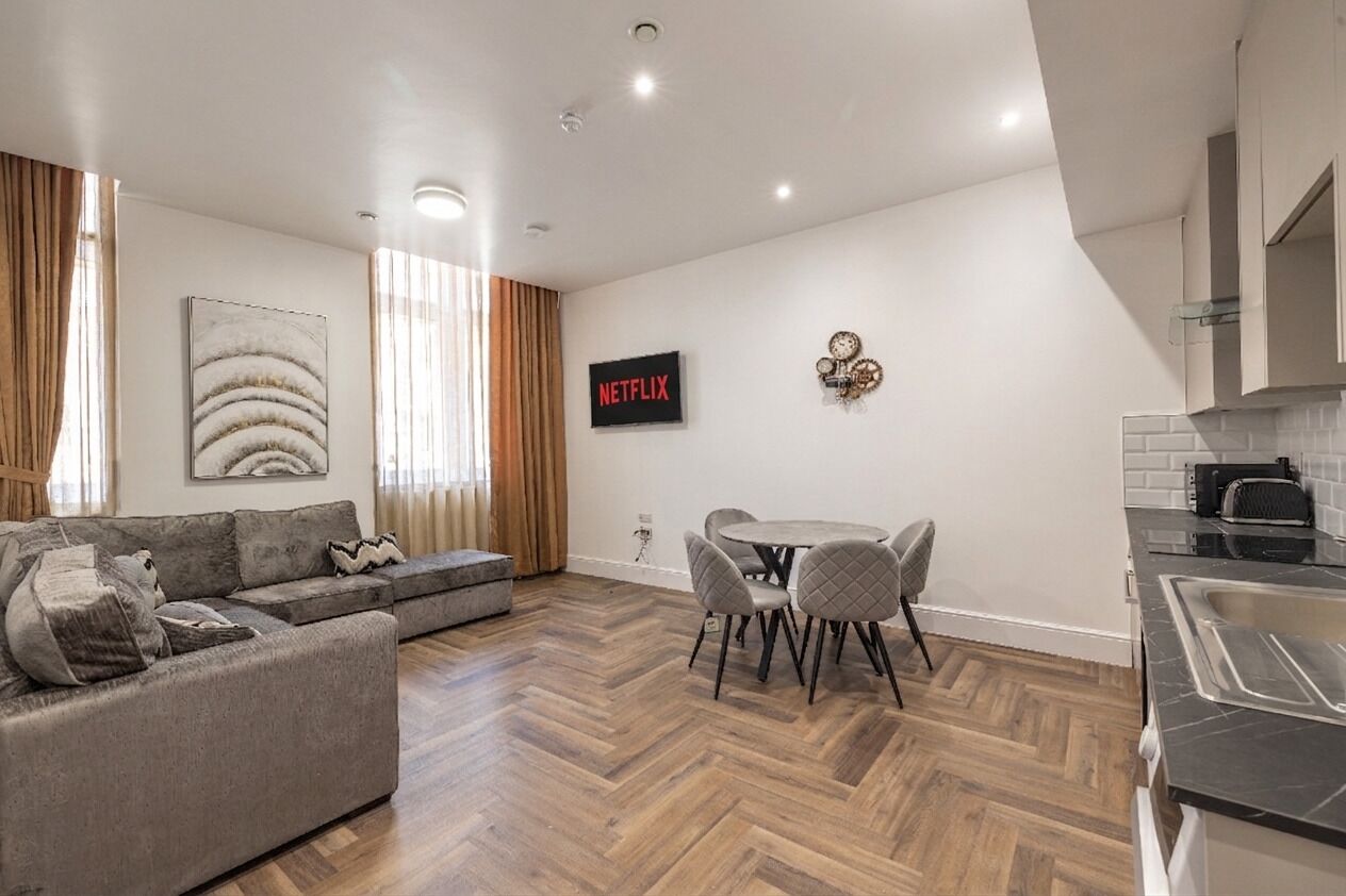 A modern living room with a sofa, a dining table with chairs, a kitchen area with a sink, and a wall-mounted Netflix sign.