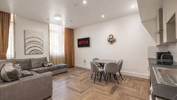 A modern living room with a sofa, a dining table with chairs, a kitchen area with a sink, and a wall-mounted Netflix sign.