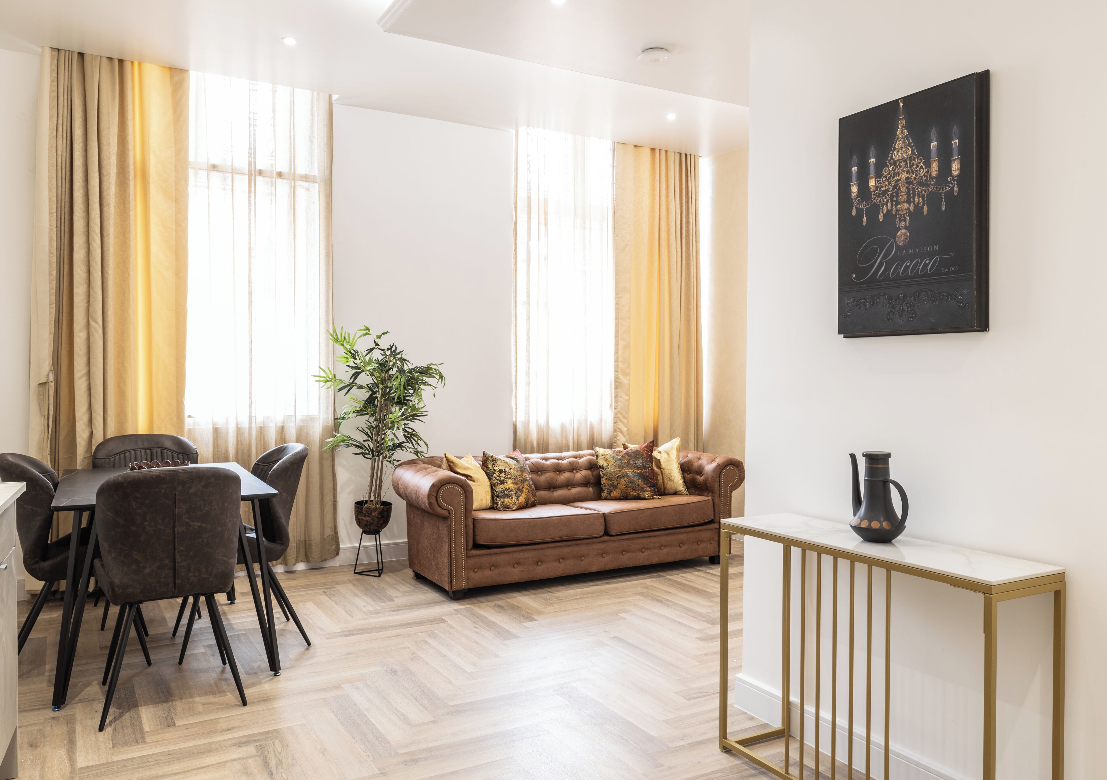 A modern living room with a sofa, dining table, chairs, a potted plant, and a wall-mounted artwork.