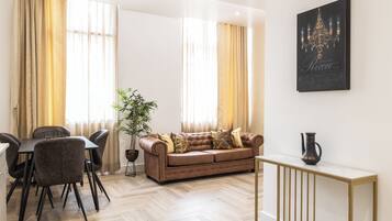 A modern living room with a sofa, dining table, chairs, a potted plant, and a wall-mounted artwork.