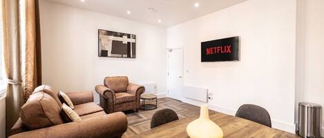 A modern living room with a sofa, armchairs, a wooden dining table, a wall-mounted TV, and a framed picture.