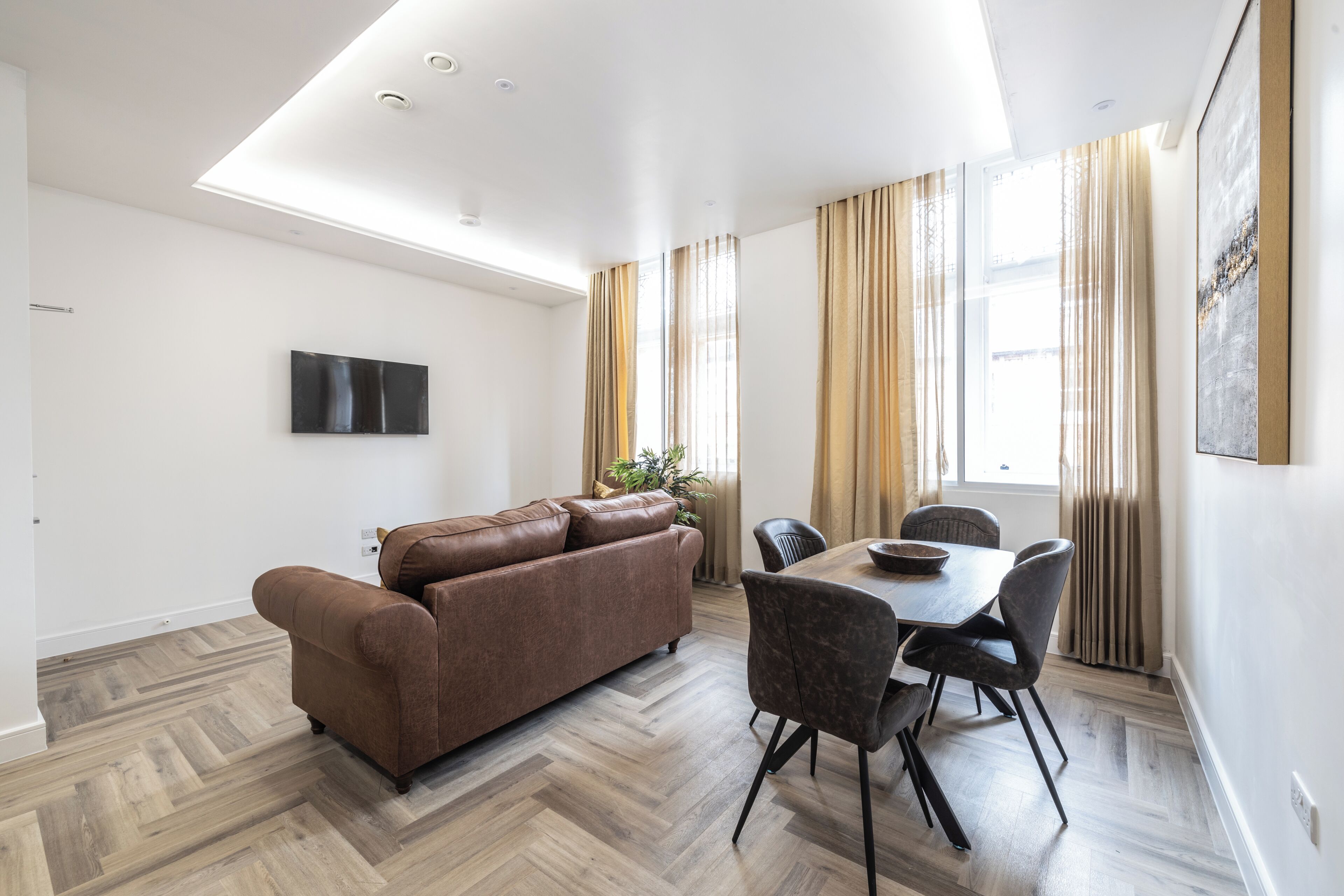 A modern living room with a brown sofa, a dining table with chairs, a flat-screen TV mounted on the wall, and a large window with curtains.