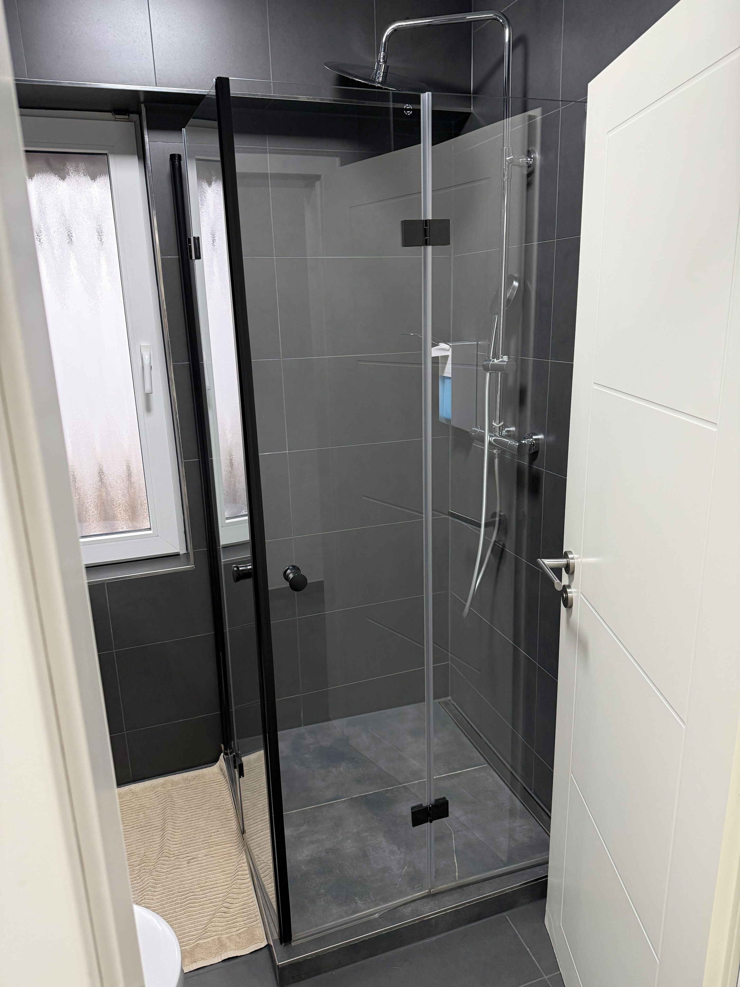 Ein modernes Badezimmer mit einer in Glas eingefassten Dusche, dunkel gefliesten Wänden und einer weißen Tür.