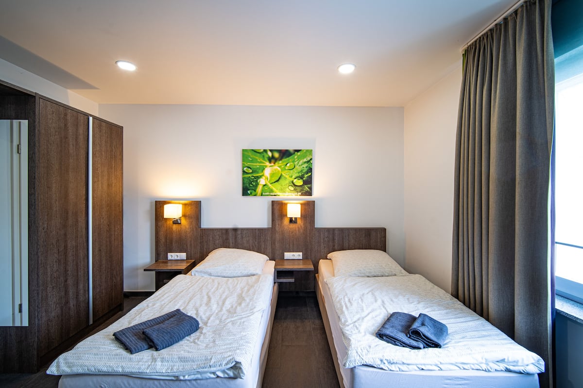 A hotel room with two single beds, a nightstand with a lamp, a wall-mounted artwork, and a window with curtains.