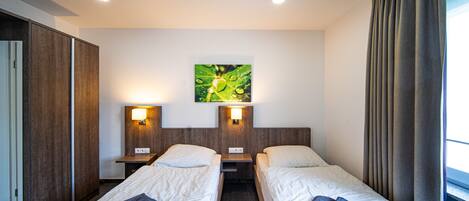 A hotel room with two single beds, a nightstand with a lamp, a wall-mounted artwork, and a window with curtains.
