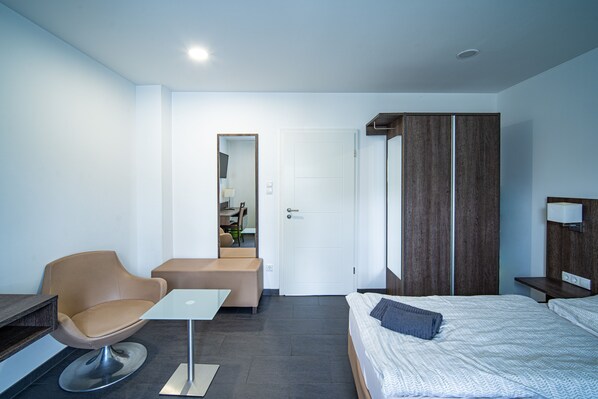 A modern hotel room with a bed, a chair, a small table, a wardrobe, and a door leading to a bathroom. - Blauer Stein - Vibe - zh2a-haus (Pulheim)
