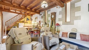 Ein gemütliches Wohnzimmer mit Holzbalkendecke, einer Couch, zwei Sesseln, einem Couchtisch und einer Treppe, die zu einer weiteren Ebene führt.