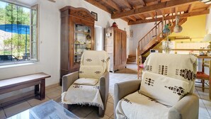 Ein Zimmer mit Holzbalken, einer Bank, zwei Sesseln mit gemusterten Decken und einer Vitrine sowie einer Treppe.