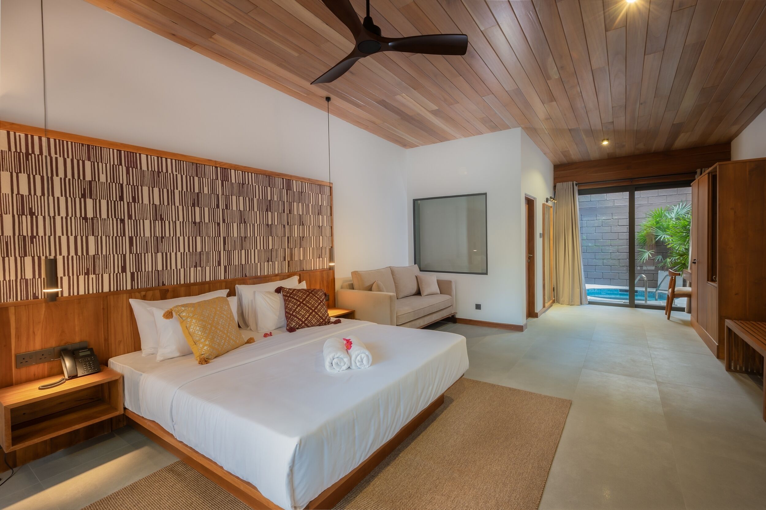 Quarto de hotel moderno com uma cama grande, um ventilador de teto, cabeceira de madeira, sofá, televisão e uma porta de vidro deslizante para uma área de piscina.