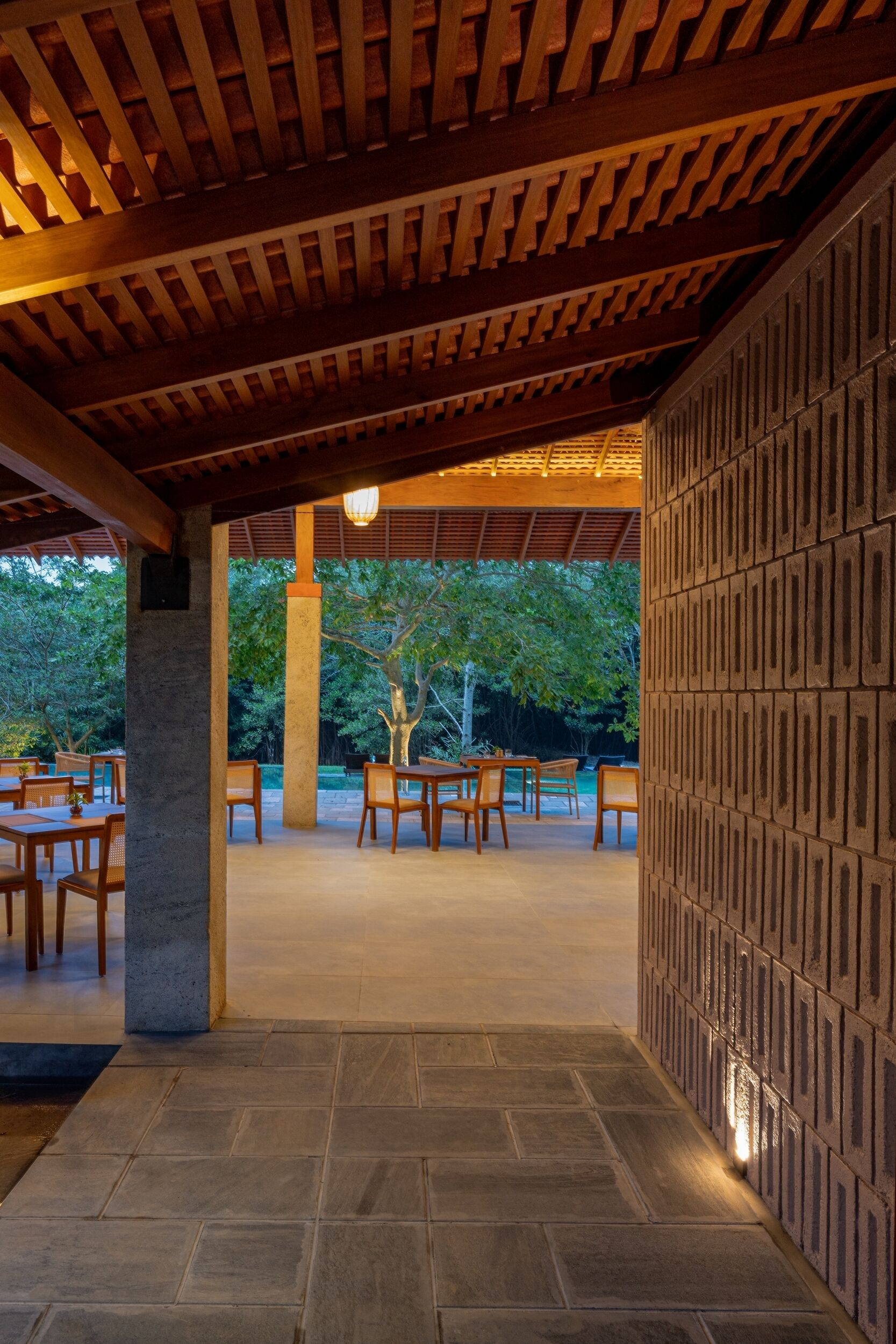 An outdoor patio area with wooden tables and chairs, a stone floor, and a wooden ceiling structure.