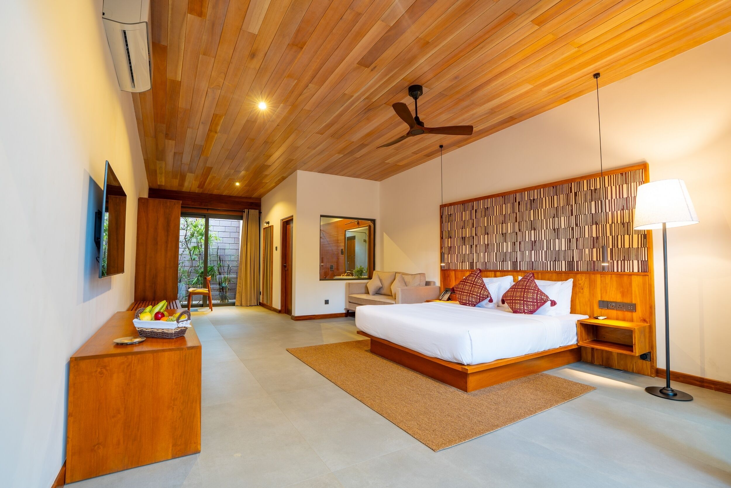 A modern hotel room with a wooden ceiling, a large bed, a desk with a fruit basket, a standing lamp, and a view of the outdoors.