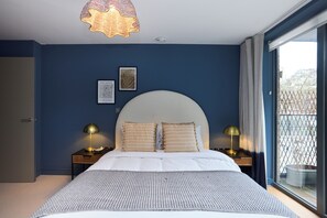 Ein Schlafzimmer mit einem großen Bett, zwei Nachttischen mit Lampen, einer Deckenleuchte und Blick ins Freie durch ein Fenster.