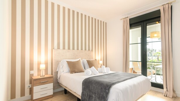 Ein ordentlich bezogenes Bett mit Kissen und Decke, ein Nachttisch mit Lampe sowie ein Fenster mit Vorhängen.