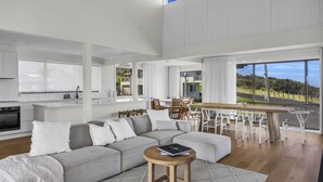 A spacious living area with a large grey sectional sofa, wooden dining table, and chairs. A kitchen with white cabinetry and a large window is visible in the background. - Fishers - Scenic Drive 35 (Bermagui)