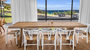 A dining area with a large wooden table and white chairs, overlooking a coastal road with parked cars and a view of the ocean. - Fishers - Scenic Drive 35 (Bermagui)