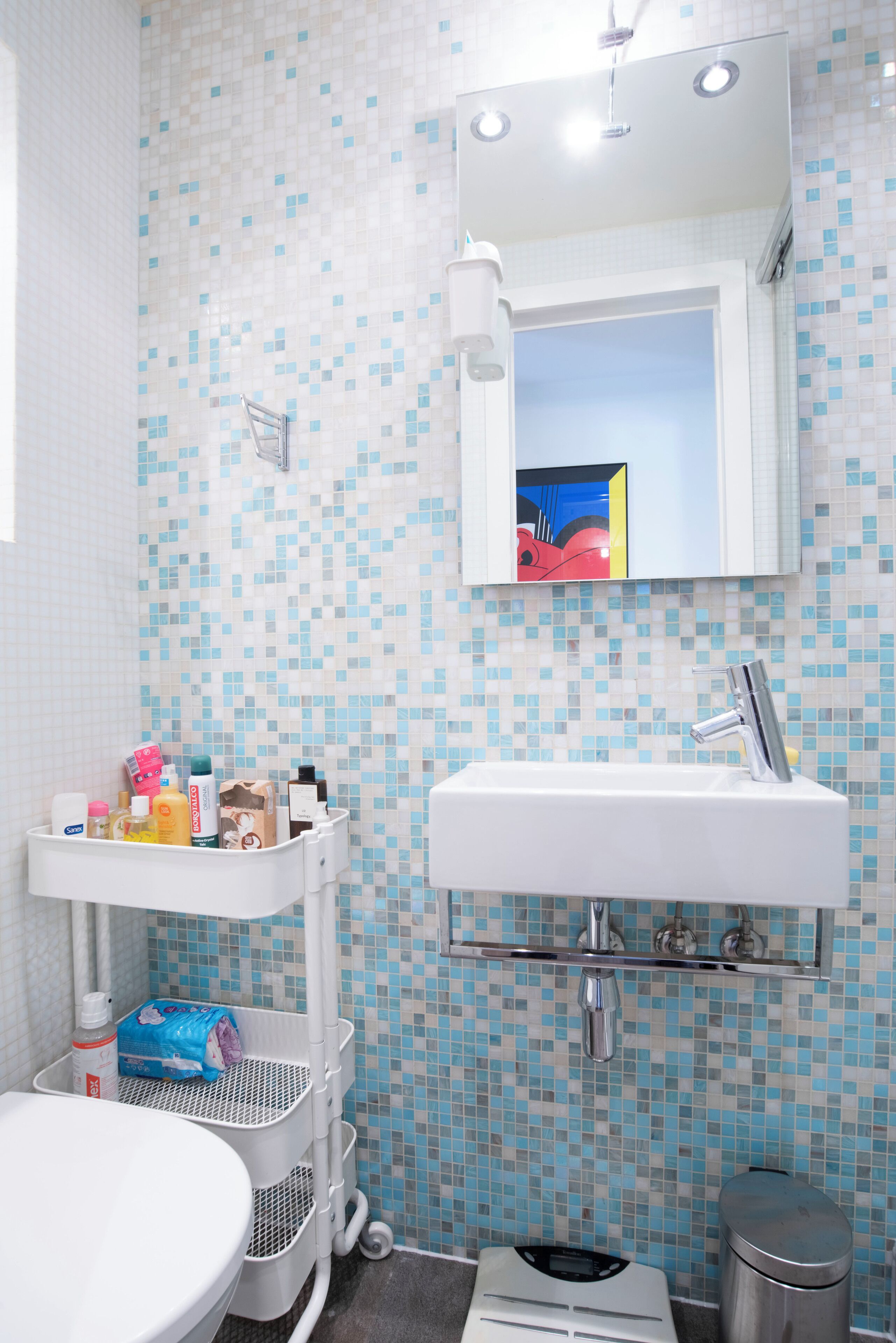 Ein Badezimmer mit einer wandförmigen Fliesenmosaik, einem weißen Waschbecken mit Spiegel darüber, einer weißen Toilette, einem Mülleimer und einem weißen Aufbewahrungswagen mit Toilettenartikeln.