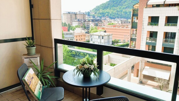 En balkon med et bord og stole, en potteplante og udsigt til bygninger og en bro.
