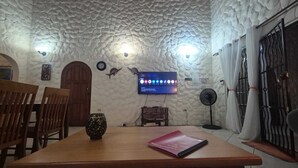 Una habitación con techo texturizado, una mesa de madera con un cuaderno, un televisor montado en la pared y un ventilador.