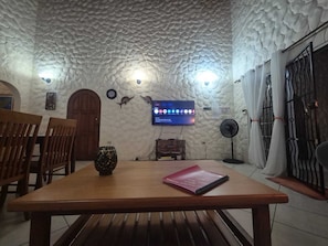Una habitación con techo texturizado, una mesa de madera con un cuaderno, un televisor montado en la pared y un ventilador.
