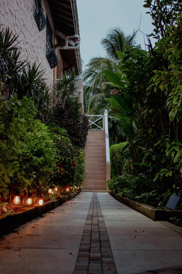 Un chemin menant à un bâtiment doté d’une entrée fermée par un portail, entouré d’une végétation luxuriante et de plantes.