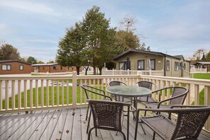 Eine Holzterrasse mit Korbstühlen und einem Glastisch, mit Blick auf eine Rasenfläche und eine Reihe von Ferienhäusern.