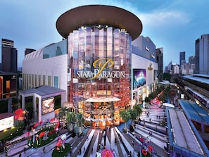 Een modern gebouw met een opvallend Siam Paragon bord, omringd door een voetgangersgebied met decoratieve bloemen en palmbomen.